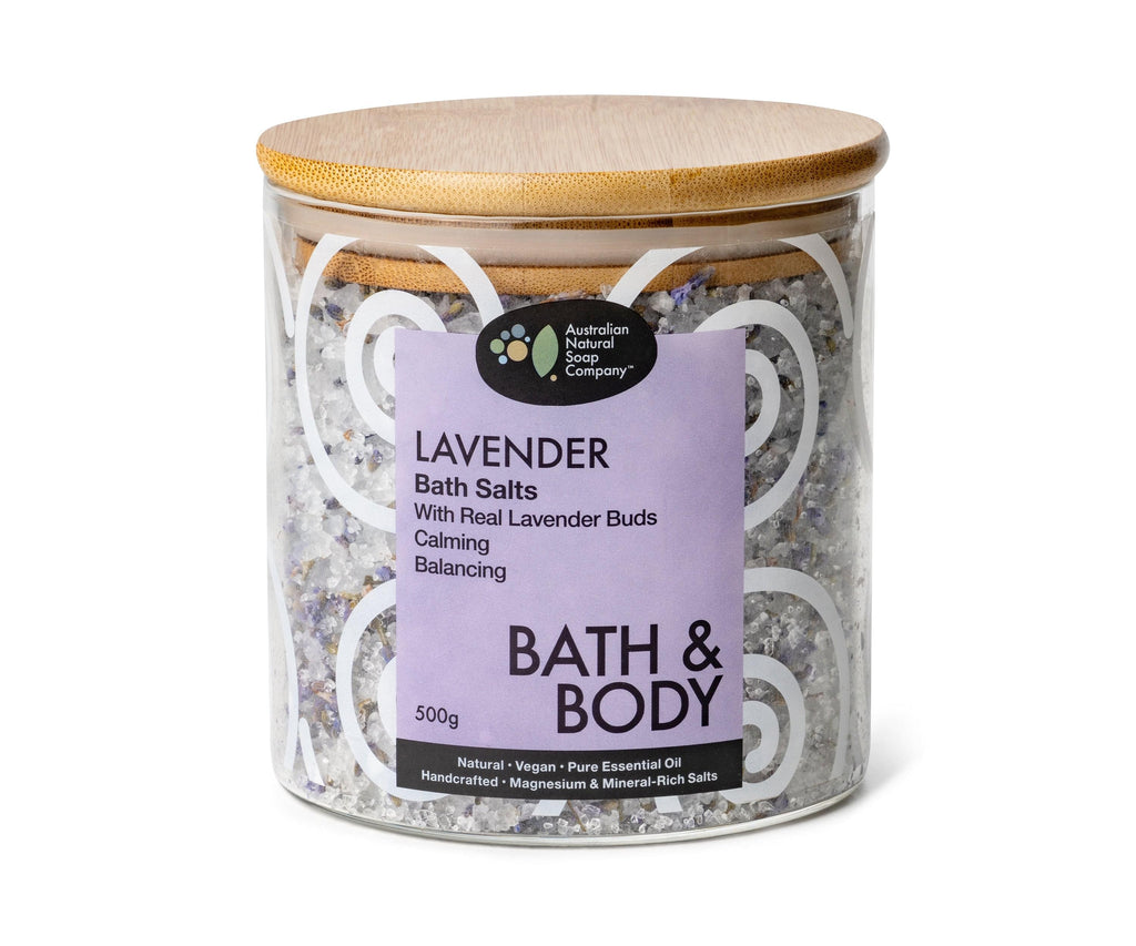 Image of a glass jar with a wooden lid. The jar is adorned with a wrap-around sticker featuring a purple color on the front and white swirling patterns around it. The sticker clearly indicates that the jar contains Lavender bath salts.