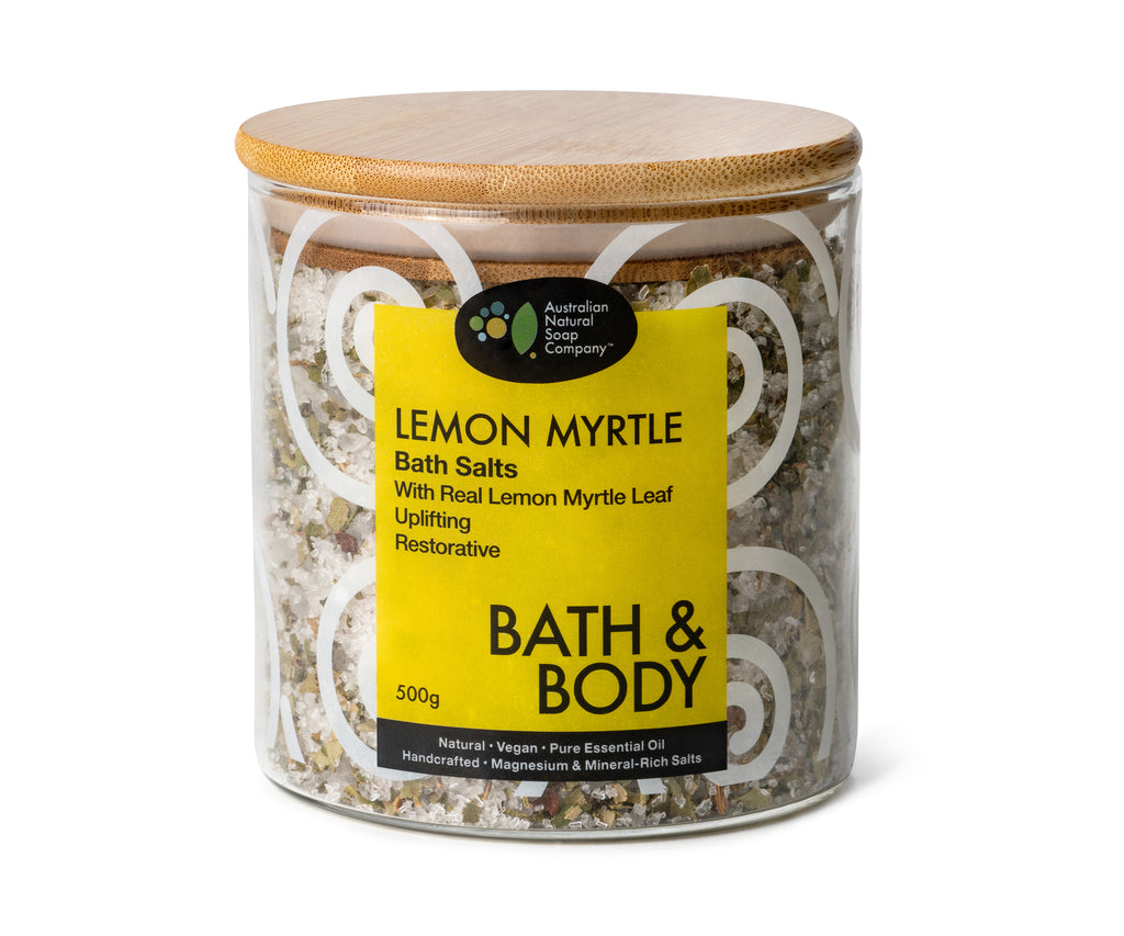 Image of a glass jar with a wooden lid. The jar is adorned with a wrap-around sticker featuring a yellow color on the front and white swirling patterns around it. The sticker clearly indicates that the jar contains Lemon Myrtle bath salts.
